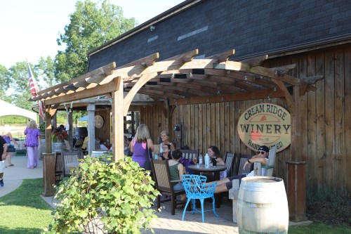 Outdoor seating area at Cream Ridge Winery, featuring wooden structures, people socializing, and a rustic backdrop.