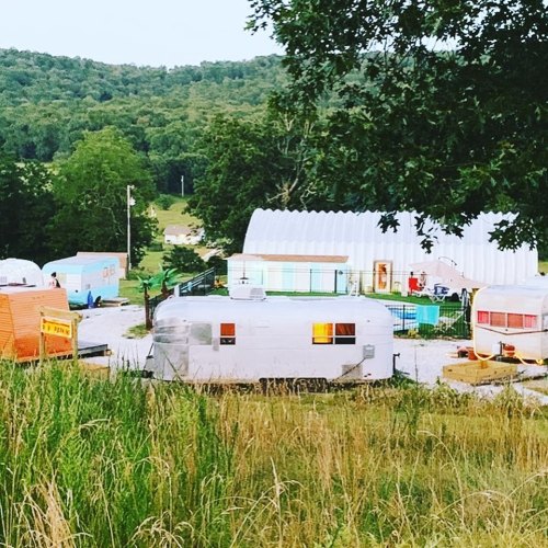 A scenic view of vintage trailers and a large building surrounded by greenery at dusk.