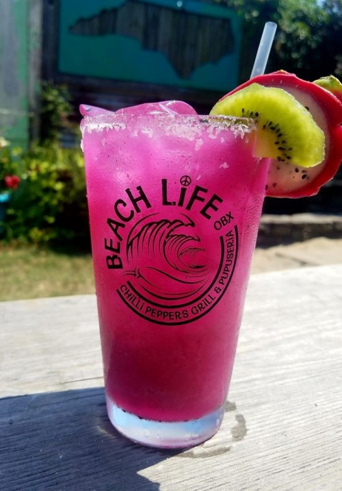 A vibrant pink cocktail in a glass with "BEACH LIFE OBX" logo, garnished with kiwi and a slice of cucumber.