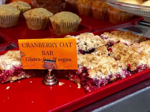 A display of cranberry oat bars labeled as gluten-free and vegan, with muffins in the background.