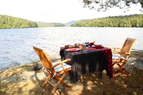 A lakeside dining setup with a table, chairs, and a scenic view of trees and water in the background.