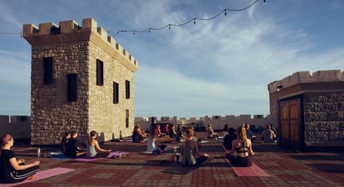 A group of people practicing yoga on a rooftop with castle-like structures and string lights in the background.