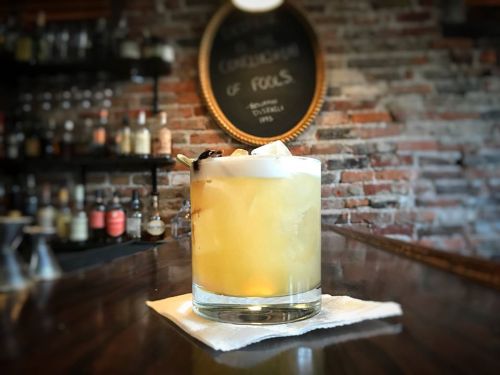 A cocktail in a glass with ice, garnished with a cherry, on a bar counter with a rustic brick wall in the background.