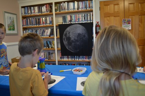 Children at a table in a library, coloring moon images with a large moon poster in the background.