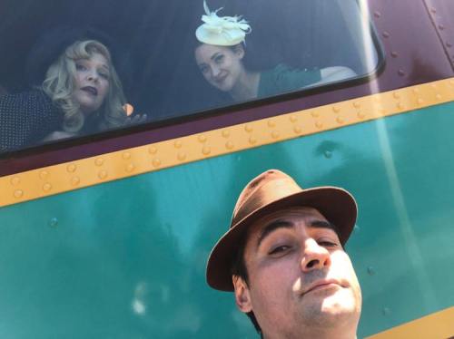 A man takes a selfie with two women in vintage attire peeking out of a train window. Bright sunlight enhances the scene.
