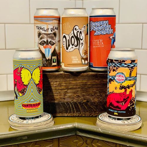 Five colorful beer cans displayed on a wooden crate, featuring unique designs and labels, against a tiled background.
