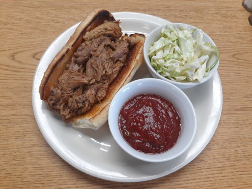 A plate with a pulled pork sandwich on a bun, a side of coleslaw, and a bowl of ketchup.