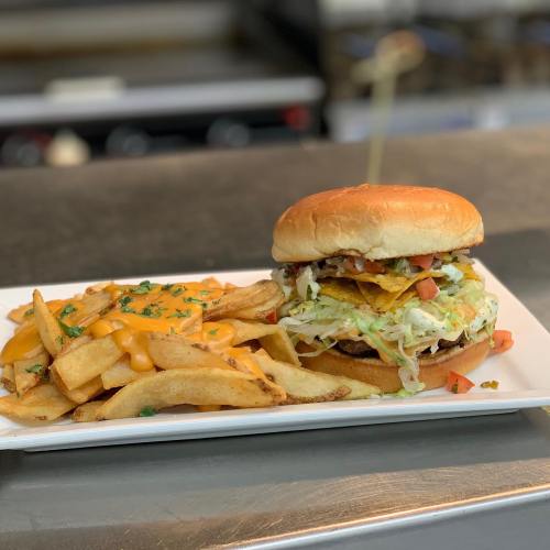 A burger topped with lettuce, tomatoes, and nacho chips, served with fries and cheese sauce on a white plate.