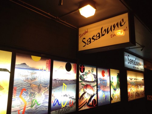 Sign for Sushi Sasabune with colorful artwork depicting waves, mountains, and seafood. Warm lighting enhances the ambiance.