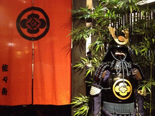A traditional samurai armor displayed next to an orange banner with a black emblem, surrounded by bamboo.