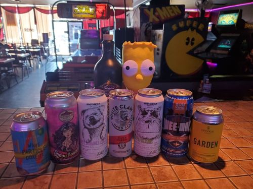 A collection of various beer cans and a bottle, with a Bart Simpson figure in a retro arcade setting.