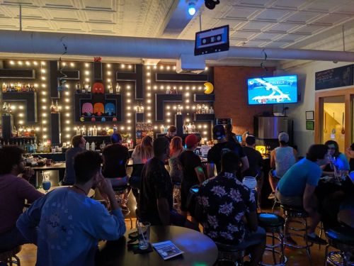 A lively bar scene with patrons enjoying drinks, neon lights, and a large screen displaying a game.