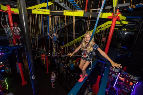 A girl balances on a high ropes course, surrounded by colorful structures and other participants in a fun indoor setting.