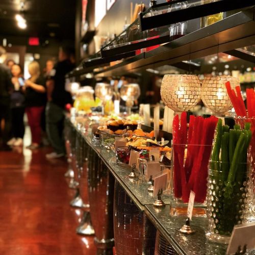 A vibrant buffet display with colorful food items and drinks, set against a stylish backdrop and a crowd in the background.