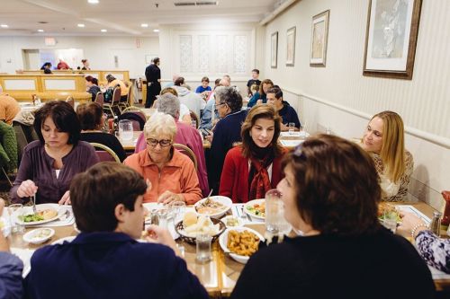 A busy restaurant scene with diverse diners enjoying meals at a large table, engaged in conversation.