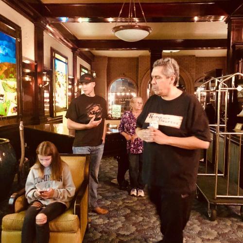A group of four people in a vintage-style lobby, with one person sitting and using a phone, others standing nearby.