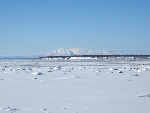 A snowy landscape with a frozen body of water and distant mountains under a clear blue sky.