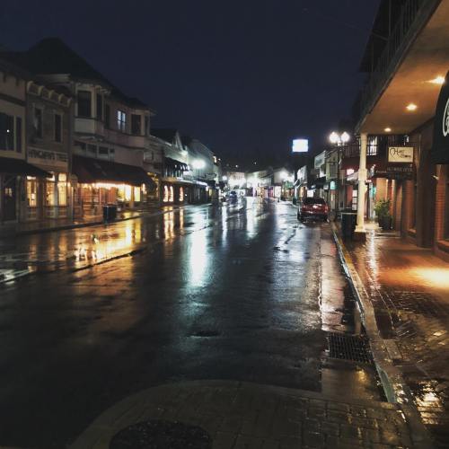 A quiet, wet street at night, reflecting lights from nearby buildings and shops.
