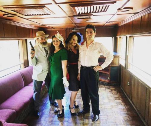 Four people dressed in vintage attire pose inside a retro train car, exuding a classic, stylish vibe.