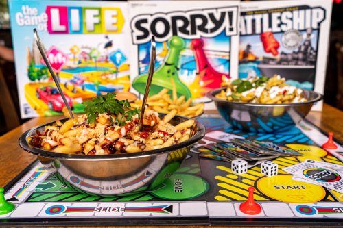 A table with two bowls of fries topped with sauces, surrounded by board games like LIFE, Sorry, and Battleship.