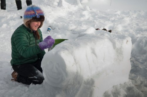 A person in a green jacket and colorful hat carves a snow sculpture in a snowy landscape.