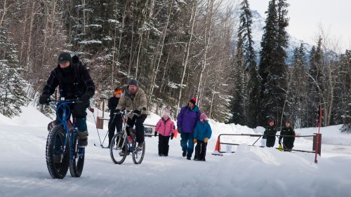 A group of people walking and biking on a snowy path surrounded by trees in a winter landscape.