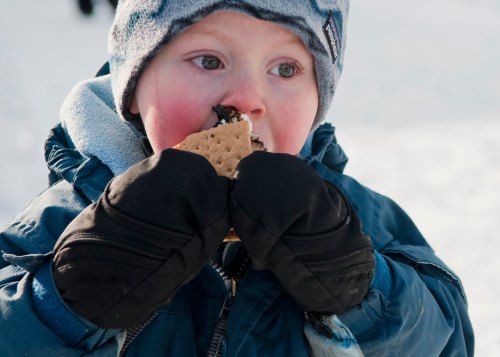 A young child in winter clothing enjoys a s'more, with chocolate and marshmallow visible, against a snowy background.