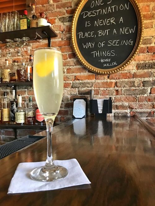 A tall cocktail glass with a lemon twist sits on a bar counter, with a brick wall and a quote in the background.