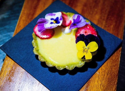 A vibrant lemon tart topped with strawberries and edible flowers, presented on a dark square plate.
