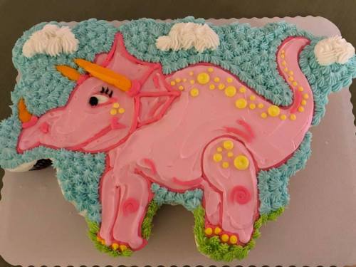 A colorful pink dinosaur cake with blue frosting, yellow accents, and decorative clouds on a silver platter.