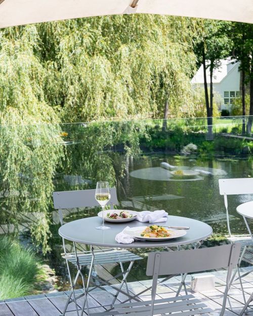 A serene outdoor dining setup by a pond, featuring a table with plates of food and a glass of wine, surrounded by greenery.