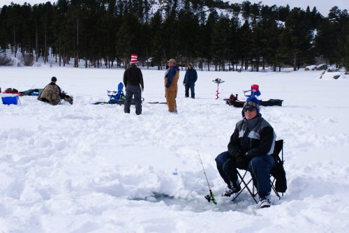 A group of people ice fishing on a snowy lake, with one person sitting on a chair by a hole in the ice.