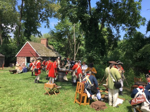 Colonial reenactors in red coats gather near a historic building, engaging in activities outdoors on a sunny day.