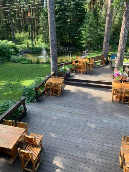 A wooden deck with tables overlooks a lush green garden and pond, surrounded by tall trees in a serene setting.