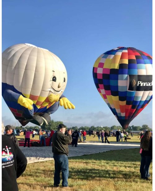 Two colorful hot air balloons, one shaped like a smiling character, are being prepared for flight at a festival.