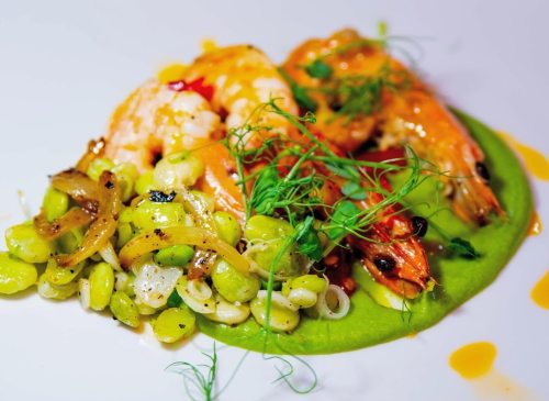 A vibrant dish featuring shrimp on a bed of green puree, accompanied by sautéed vegetables and garnished with herbs.