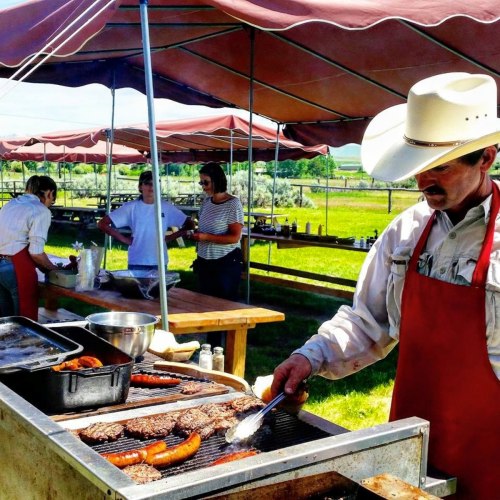 A man in a cowboy hat grills sausages and burgers at an outdoor event, with people preparing food in the background.