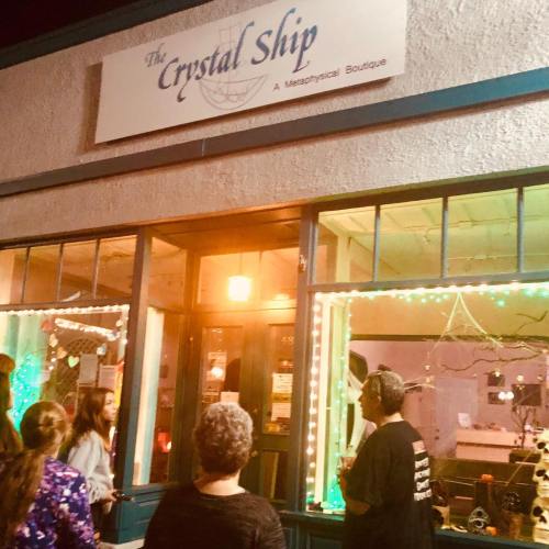 A group of people stands outside "The Crystal Ship," a metaphysical boutique, adorned with colorful lights.