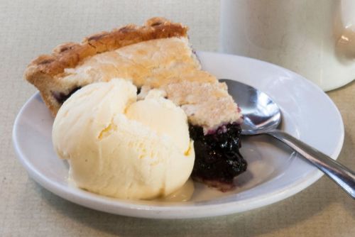 A slice of blueberry pie with a scoop of vanilla ice cream on a white plate, accompanied by a spoon.
