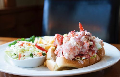 A lobster roll topped with creamy lobster salad, served with coleslaw and fries on a white plate.