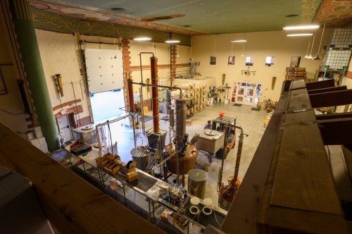 A spacious distillery interior with large equipment, barrels, and bright lighting, viewed from an upper level.