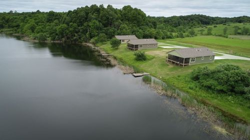 Aerial view of two houses by a calm lake, surrounded by lush greenery and trees under a cloudy sky.
