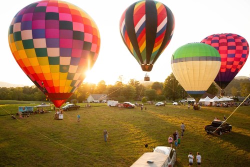 Colorful hot air balloons ascend into a sunset sky over a grassy field, with people and vehicles below.