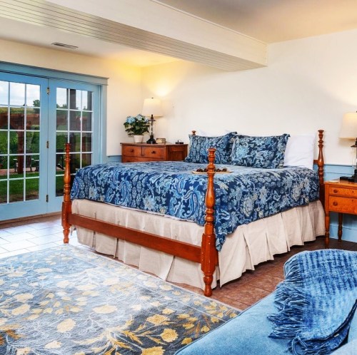Cozy bedroom with a blue floral bedspread, wooden furniture, and large windows overlooking a garden.