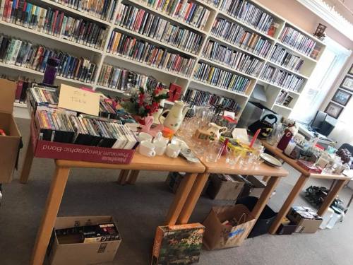 A room filled with tables displaying various items for sale, including DVDs, dishes, and boxes, in front of a library shelf.