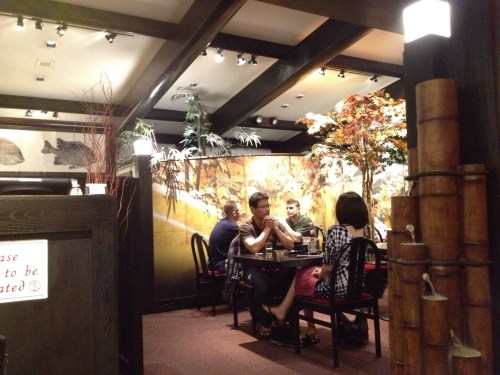 A cozy restaurant interior with four people seated at a table, surrounded by decorative plants and a scenic mural.