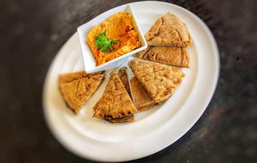 A plate with pita bread triangles and a bowl of orange hummus, garnished with a sprig of parsley.