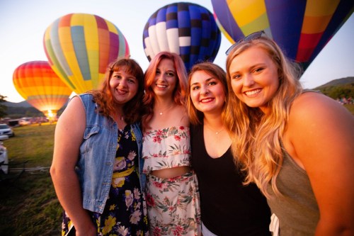 Four friends smile together in front of colorful hot air balloons at sunset.