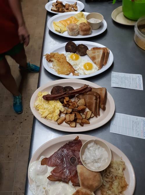 A variety of breakfast plates featuring eggs, bacon, sausage, hash browns, toast, and biscuits on a table.