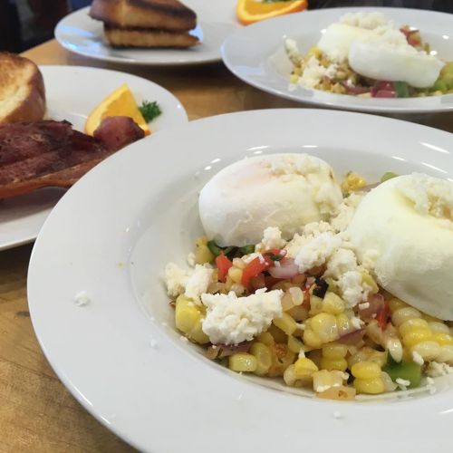 A plate of corn salad topped with poached eggs and feta, with bacon and toast in the background.